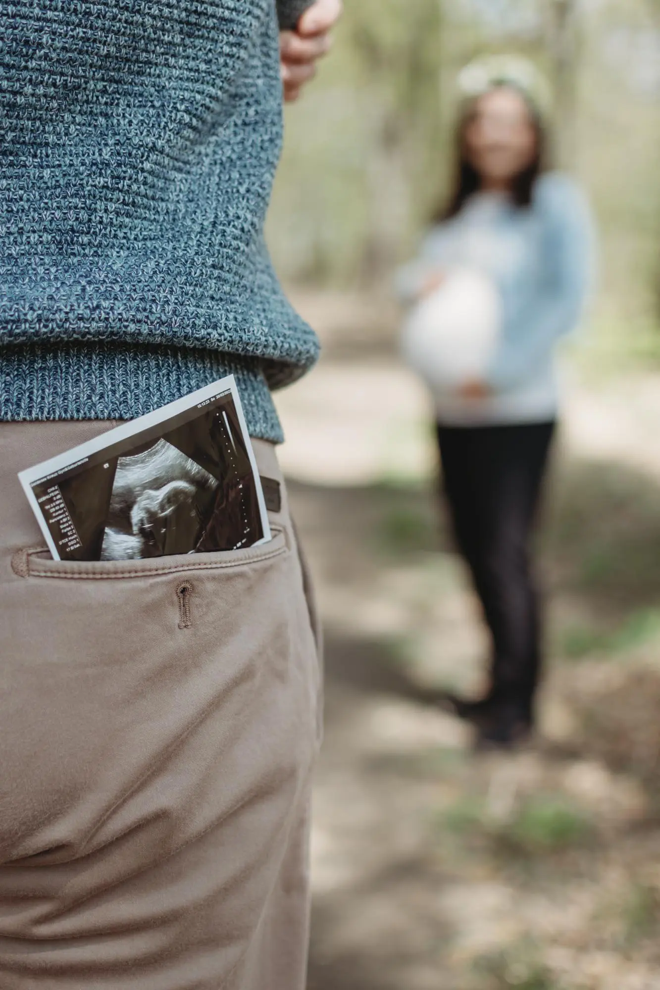 Babybauchshooting: Papa hat Ultraschallbild in Hosentasche und Mama steht im Hintergrund