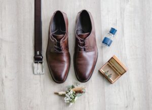 Aufnahme der Bräutigam-Accessoires. Braune Lederschuhe, Manschettenknöpfe, Ansteckblume, Parfum und Gürtel sind wunderschön vorbereitet und in einer Aufnahme von oben festgehalten