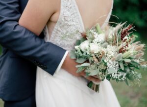 Nahaufnahme vom Brautstrauß - der Bräutigam hält den Brautstrauß hinter dem Rücken der Braut während sie ihn umarmt