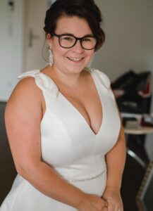 Braut beim Getting Ready. Sie strahlt in die Kamera und hat ein wunderschönes weißes Kleid mit einem Straßgürtel und Schleifen an den Schultern an. Dazu wunderschöne Glitzer-Ohrringe