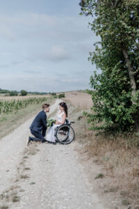 Der Bräutigam kniet vor seiner Braut im Rollstuhl auf einem Feldweg vor ihrem Lieblingsbaum und die beiden strahlen sich verliebt an