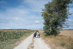 Der Bräutigam kniet vor seiner Braut im Rollstuhl auf einem Feldweg vor ihrem Lieblingsbaum und die beiden küssen sich