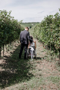 Braut und Bräutigam stehen zwischen Weinreben - Aufnahme von hinten und man sieht den geschmückten Rollstuhl