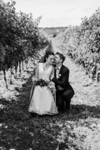 Braut und Bräutigam sind zwischen den Weinreben. Der Bräutigam kniet neben seiner Braut und küsst sie auf die Wange