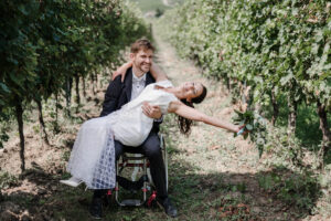 Der Bräutigam sitzt im Rollstuhl seiner Braut. Er hat sie auf seiner Schoß und sie hebt strahlend die Arme nach hinten