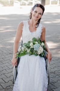 Braut im Rollstuhl mit wunderschönen Brautstrauß am Schoss lacht in die Kamera bei der Ankunft bei der Trauung