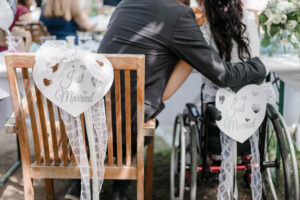Blick von hinten auf das Brautpaar, dass beim Brauttisch sitzt. Man sieht wie sich die beiden umarmen und die beiden geschmückten Stühle