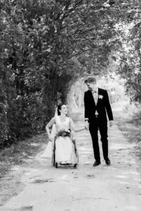 Braut und Bräutigam gehen einen Waldweg entlang. Die Braut im Rollstuhl blickt ganz verliebt zu ihrem Ehemann. Schwarz/weiß Aufnahme