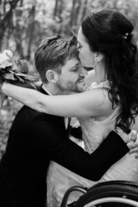 Der Bräutigam kniet vor seiner Braut im Rollstuhl. Sie legt die Arme um seinen Hals und gibt ihm einen Kuss auf die Stirn