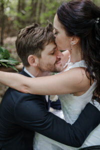 Der Bräutigam kniet vor seiner Braut im Rollstuhl. Sie legt die Arme um seinen Hals und gibt ihm einen Kuss auf die Stirn