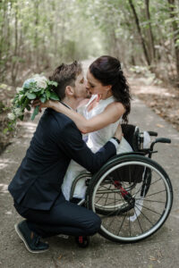 Der Bräutigam kniet vor seiner Braut im Rollstuhl. Sie legt die Arme um seinen Hals und die beiden küssen sich