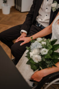 CloseUp auf die Hände des Brautpaares während der Trauung. Braut und Bräutigam haben die Hand am Schoß vom Bräutigam. Der Brautstrauß liegt auf dem Schoß der Braut im Rollstuhl