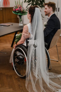 Aufnahme von schräg hinten auf das Brautpaar. Man sieht wie der Bräutigam zu seiner Braut sieht und dabei den Arm um ihre Schulter liegen hat