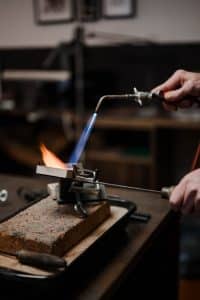 Der Goldschmied schmilzt ein Stück Metal für die Weiterverarbeitung. Man sieht die Flamme eines Bunsenbrenners in einer Schmelzform