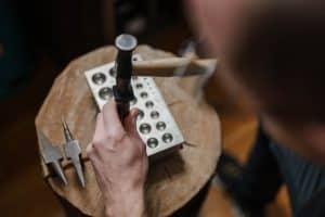 Nahaufnahme des Goldschmieds beim Formen einer Rundung von eines Metalstücks. Hierfür wird über die Schulter des Goldschmieds fotografiert und man sieht wie er mit Hammer und Metallstock das Metall in eine Form drückt