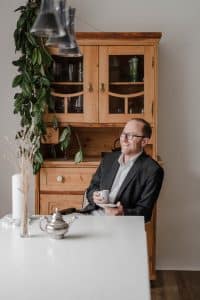 Portrait des Goldschmieds im Rahmen einer Businessreportage in Wien. Er sitzt mit einer Tasse Kaffee am Esszimmertisch und schaut lachend aus dem Terrassenfenster.