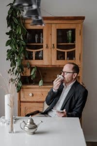 Portrait des Goldschmieds im Rahmen einer Businessreportage in Wien. Er sitzt mit einer Tasse Kaffee am Esszimmertisch, schaut lachend aus dem Terrassenfenster und trinkt.