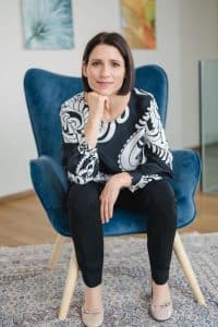 Portrait einer Anwältin im Rahmen einer Businessreportage in Wien. Sie sitzt in einer lichtdurchfluteten Wohnung auf einem blauen Stuhl mit Holzbeinen, lehnt sich ein Stück nach vorne und stützt sich mit dem Kinn auf ihrer rechten Hand ab. Dabei lächelt sie leicht in die Kamera