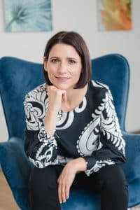 Nahaufnahme einer Anwältin im Rahmen einer Businessreportage in Wien. Sie sitzt in einer lichtdurchfluteten Wohnung auf einem blauen Stuhl mit Holzbeinen, lehnt sich ein Stück nach vorne und stützt sich mit dem Kinn auf ihrer rechten Hand ab. Dabei lächelt sie sympatisch in die Kamera und strahlt Stärke für ihre Mandant*innen aus