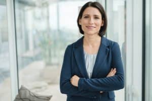 Portrait einer Anwältin im Rahmen einer Businessreportage in Wien. Sie steht an einer Fensterfront und lehnt sich ganz leicht an das Fenster. Dabei schaut sie mit leicht verschränkten Armen direkt in die Kamera und lächelt dabei