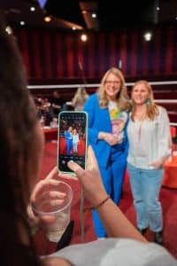 Buinessreportage_Veranstaltungsreporte: Live Video Podcast von Corinna Hintenberger mit Stefan Verra im socialhub Wien. Corinna Hintenberger posiert mit einer Zuschauerin im Studio wo der Podcast aufgezeichnet wurde für ein Handyfoto. Auf diesem Bild sieht man das Foto auf dem Handy und der Hintergrund ist unscharf